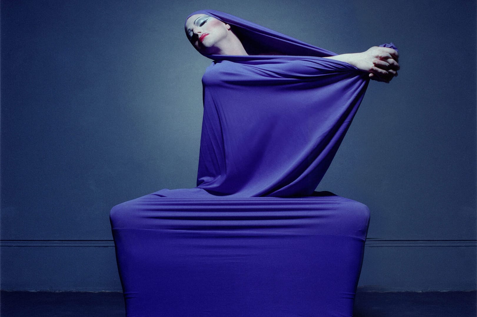 Richard Move wears a tight sheer dress of a rich dark blue, with only their face, hands, and feet visible. Their legs are open wide, only the balls of the feet on the floor, and they extend their gripped hands to the left as they dramatically turn their head to the right. Fans of modern dance will recognize that this image is a recreation of the dance solo "Lamentations" by Martha Graham.