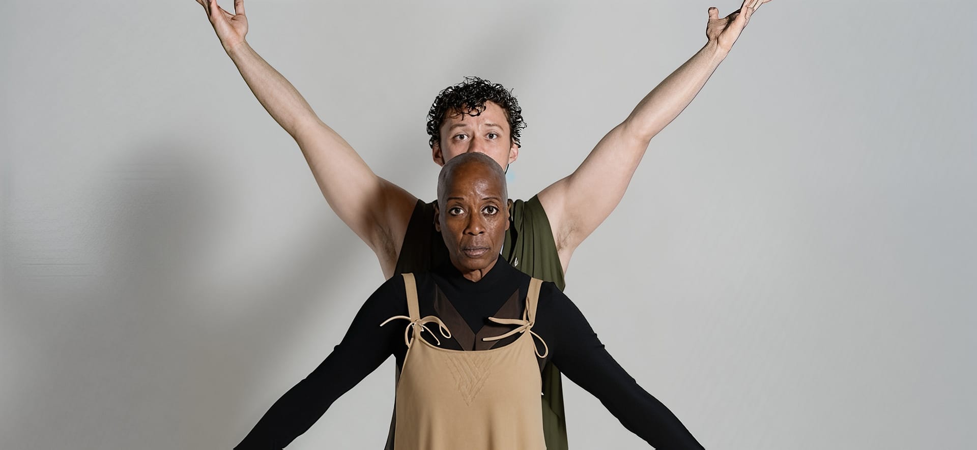 Rhonda Moore stands with her arms outstretched in a low “V” position looking directly at the camera. Ben Grinberg stands behind her with his arms outstretched in a high “V” position looking at the camera. Grinberg wears a green sleeveless top, and Moore is wearing a longsleeve black button-down under a light-brown jumpsuit. They stand in front of a gray backdrop.