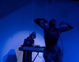 Vyette Tiya (she/her) smiles with her eyes closed and head tilted upwards, arms lofting upwards as she descends downwards. She wears baggy pants and a textured crop top, and is washed in a dark blue light. Two white chains hang in the background above her head. To her left is Daniel Sohn with a mixing board and laptop, focused on mixing for his music. He is wearing a baseball cap, t-shirt, and pants.