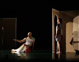 (Left to Right): Burr Johnson, Savannah Gaillard. A bald man with a beard sits on the floor, he wears what looks like a loose skirt or dress, and gazes at his right arm which is gently extended horizontally from his side. Nearby, a woman with dark hair wearing shorts stands in profile in front of a wall made out of tatami, a small doorway in front of her.