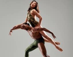 The image features a powerful duet from Complexions Contemporary Ballet. A male dancer, dressed in shimmering green pants, supports a female dancer in a gold costume as she elegantly balances on his shoulders, demonstrating strength, balance, and grace in their contemporary ballet movement.