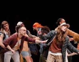A group of standing performers lean towards one another while gazing intently towards an unknown subject. One performer, wearing a maroon jacket and glasses, grips the body of the jacket of a performer with long hair wearing a baseball cap. Their facial expressions evoke intensity, but their bodies appear to be frozen in time.