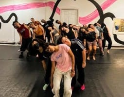 A large group of dancers stand closely together in a studio, their bodies tilted in unison with heads falling dramatically to one side, creating a strong visual wave across the room. They are arranged in staggered rows that draw the eye from front to back, emphasizing their collective rhythm and connection. The black floor grounds the image, while the wall behind them bursts with bold, curving black outlines and sweeping pink brushstrokes, giving the space an energetic, almost theatrical backdrop.