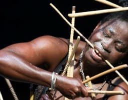 With eyes closed, Nadia Beugré holds a stick in her mouth and tilts her head to the left. More wooden sticks frame her face. She is sweaty, her right arm strong and flexed.
