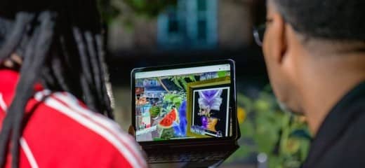 Two people look at an ipad with virtual reality images. Their backs are facing the camera and the person on the left wears a green hat, long locs, and a red shirt. The person on the right wears a black shirt and glasses.