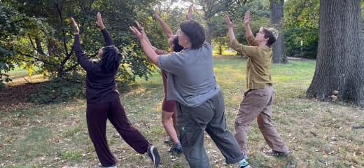a group of four individuals stand together with arms lifted overhead making a clapping gesture with their hands. They are outside in the grass, surrounded by trees and other greenery. Their outfits are earth tones - browns, greens, and shades of grey.