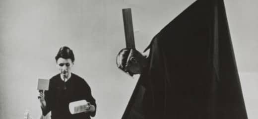 A black and white photo of Lucia Dlugoszewski kneeling on the floor holding a cylinder and a box shaped item in either hand. in front of her sits a pile of wooden objects and instruments. beside her, Hawkins, draped in a black cape-like garment which reveals only his head, tilts toward Dlugoszewski and creates a triangular shape with the garment.