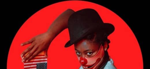 There is a black grassy horizon line and a mostly full red moon rising. In the circle of the moon is a young black woman in a short sleeve white shirt and a black vest. Her Right arm is raised so her hands can frame above and below a small upside down american flag. She wears a circus style top hat and a mask over the bottom half of her face that is a clown's wide smile.