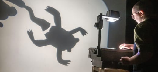 Madeline Shuron, wearing denim overalls and glasses, uses a vintage overhead projector to share the cut-out image of a frog losing its tail. We see the silhouette of this large-scale creature.
