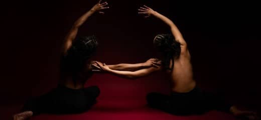 Two dancers sit on a red floor in front of a black backdrop with their hair braided into updos with cornrows. They are topless with their bare backs facing the camera, wearing black leggings. They mirror each other, leaning toward one another with one hand outstretched, resting on the other’s shoulder. The other arm arcs overhead, also in the direction of the other.